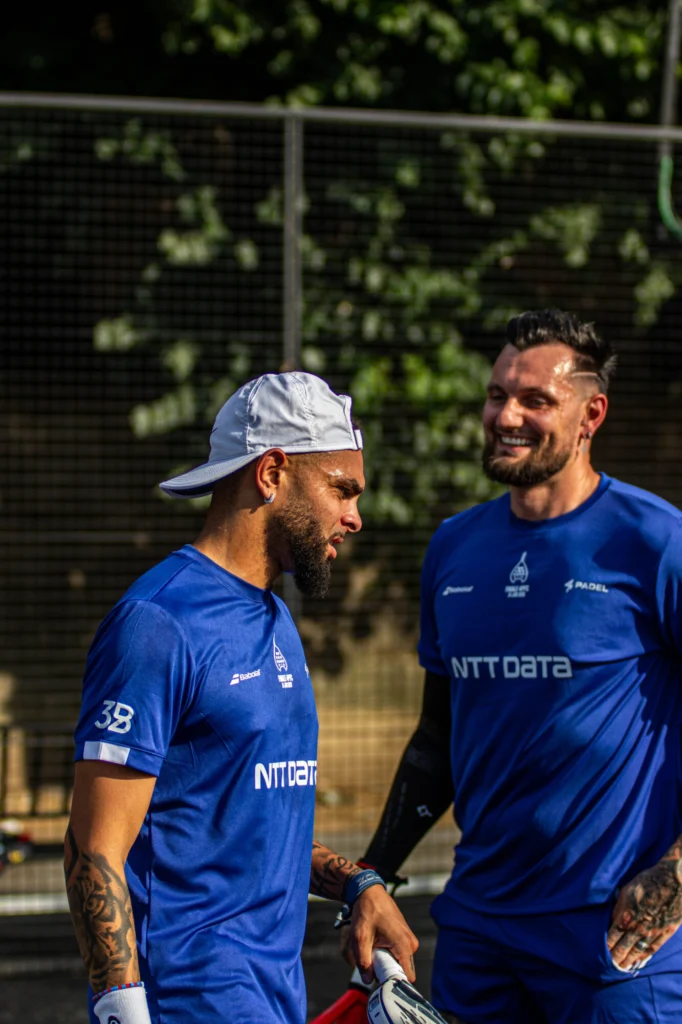 369A7788 | Padel Magazine Two men in blue athletic shirts stand together outdoors, smiling during a padel game, with a fence and greenery in the background.