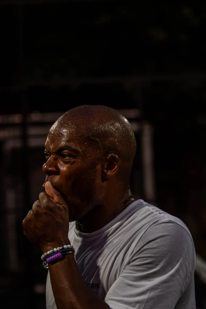 Close-up of a sweating man with a shaved head and gray shirt, resting his chin on his hand in a dark setting.