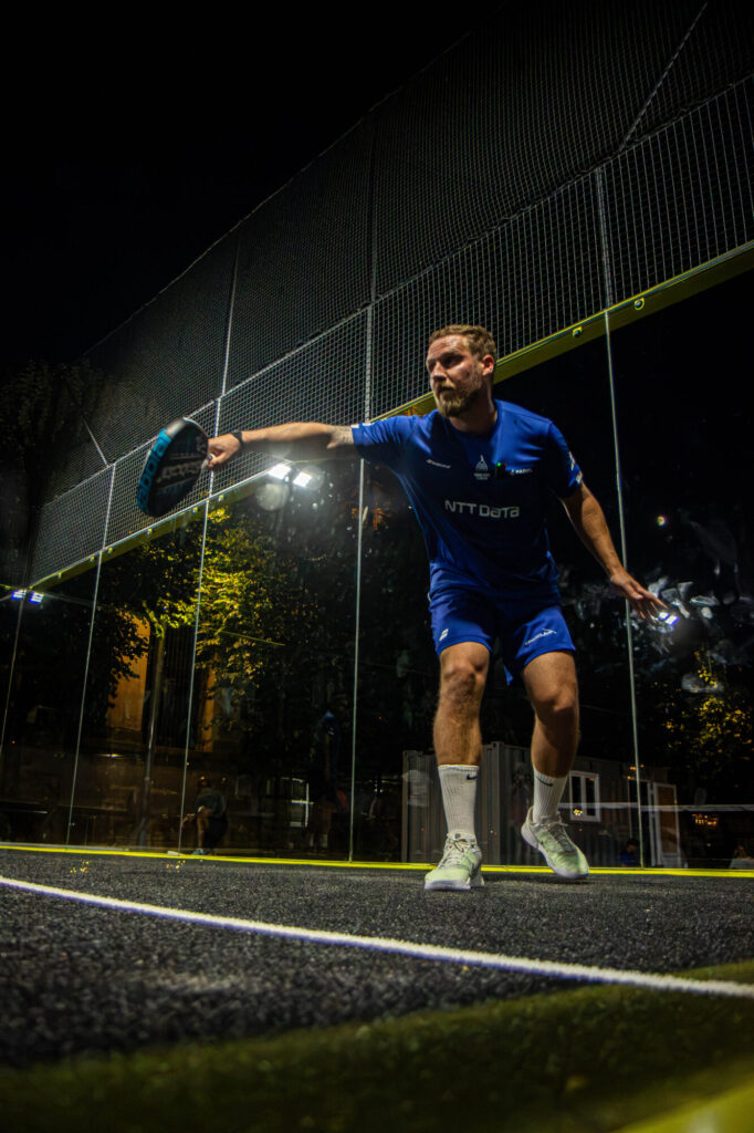 369A8500 | Padel Magazine A man in a blue sports kit swinging a paddle on a glass-walled padel court at night, preparing to hit the ball.
