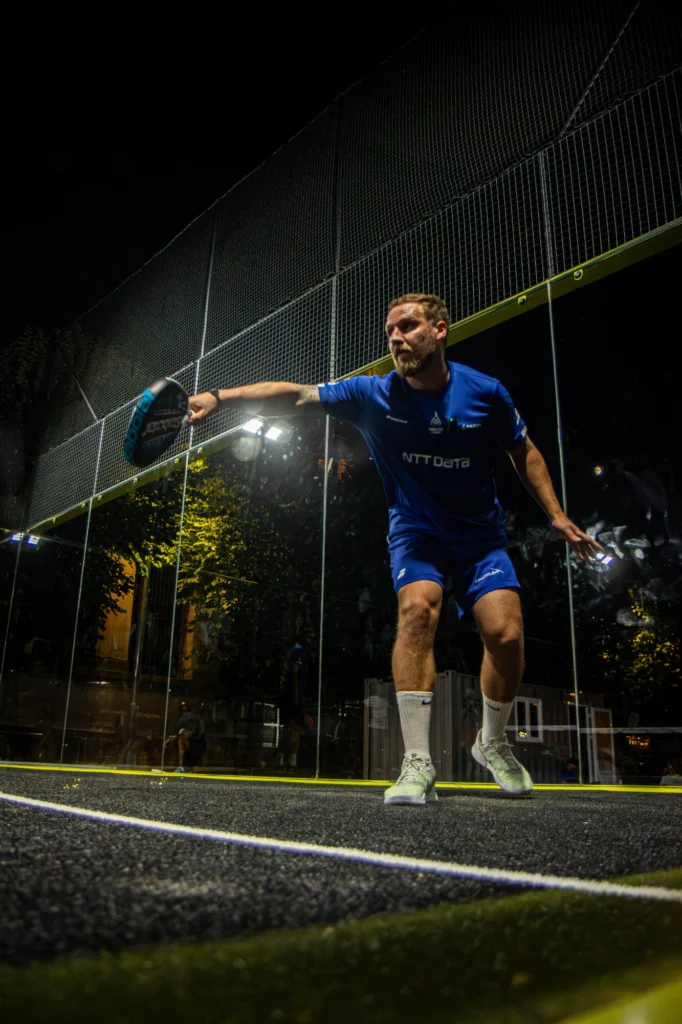 A man in a blue sports kit swinging a paddle on a glass-walled padel court at night, preparing to hit the ball.