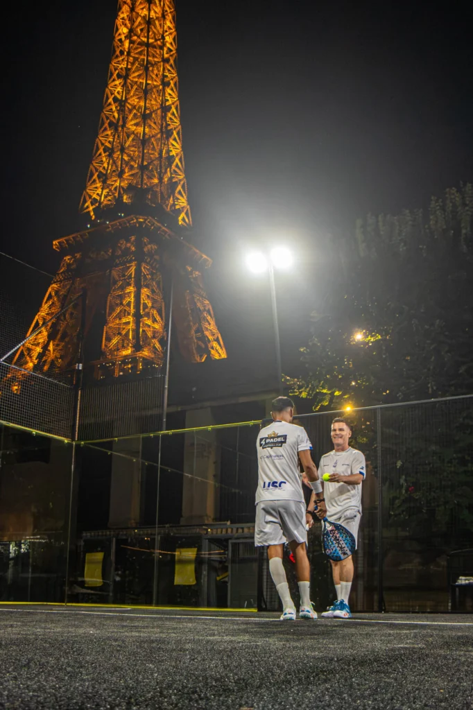Two men in white padel outfits stand on a court at night, chatting with a racket and ball in hand, with a tall orange lattice tower in the background.
