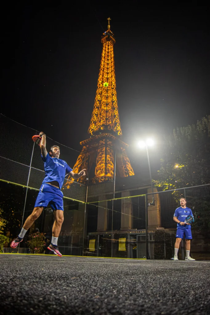 Nighttime padel match on a glass-walled court, a player in blue leaps to hit the ball with a paddle as a lit lattice tower glows in the background.