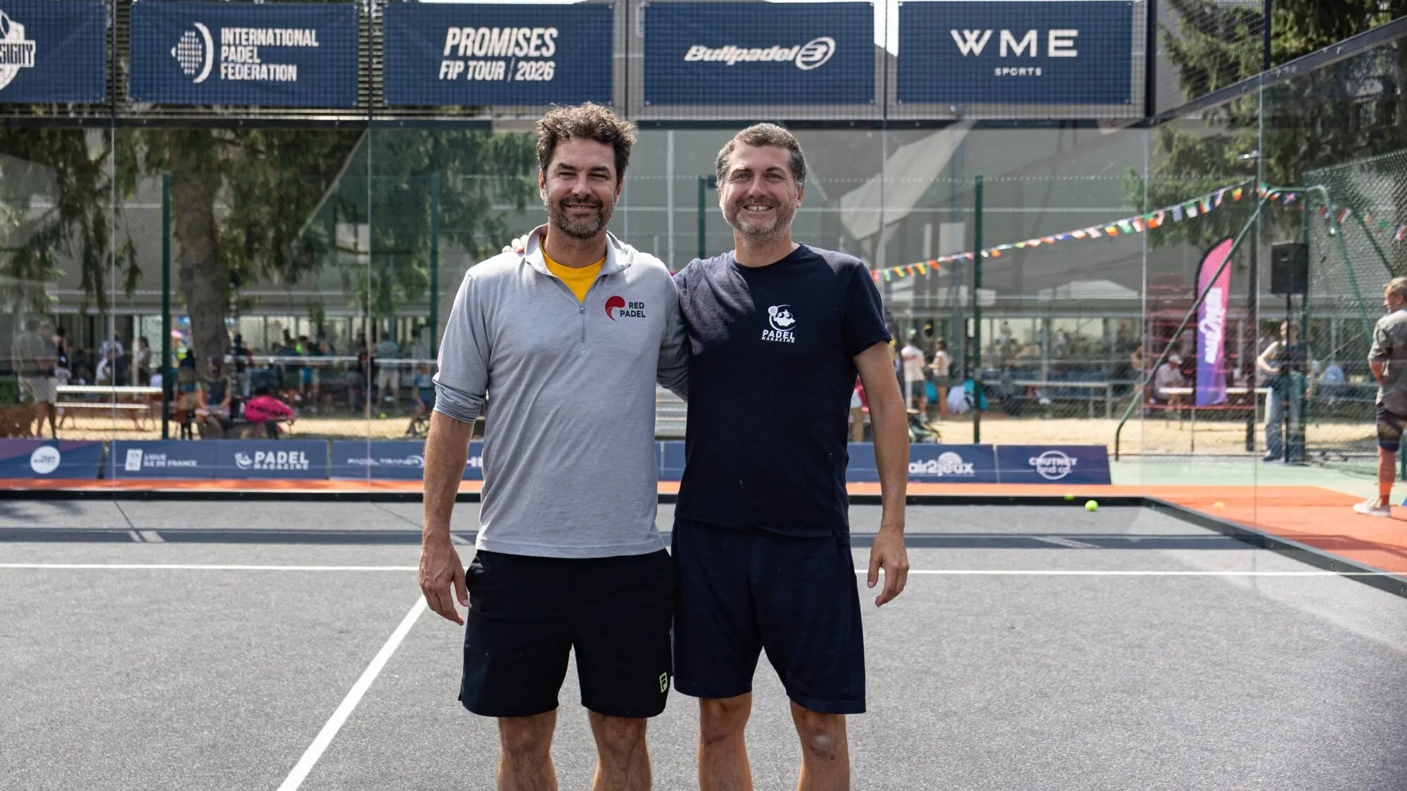 Two men stand together on a padel court, smiling, with banners for the International Padel Federation in the background.