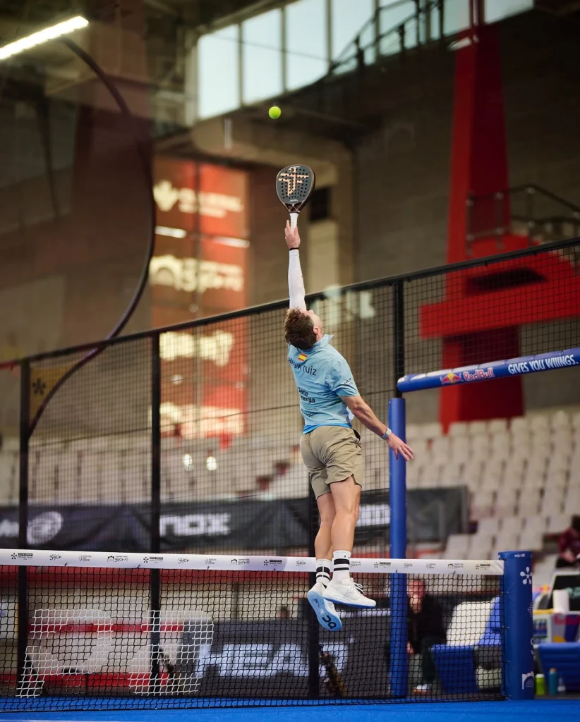 Padel Ein Spieler in einem hellblauen Hemd springt über das Netz, um einen gelben Ball mit einem Schläger zu schlagen. Die Aufnahme zeigt eine Szene aus einer Indoor-Arena am Spielfeldrand.
