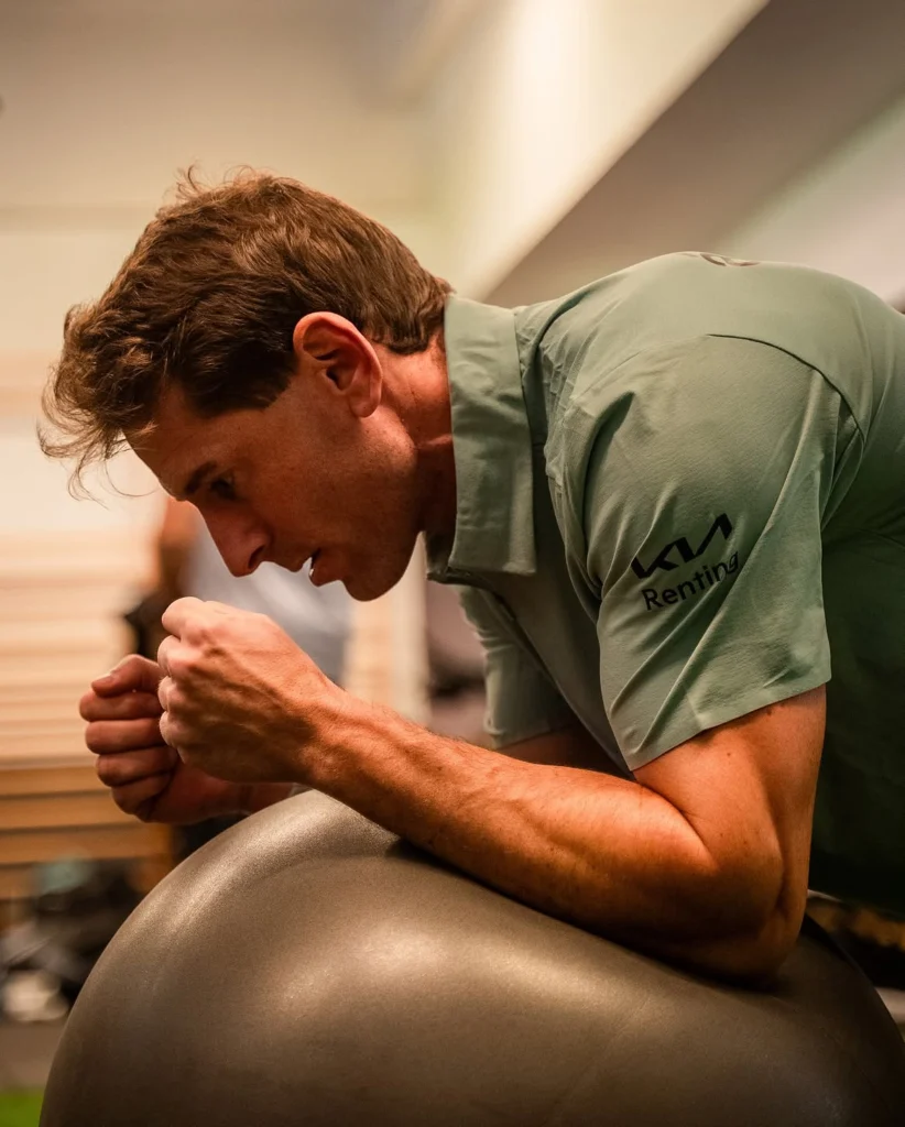 Ein Mann in einem grünen Polohemd beugt sich über einen großen Gymnastikball und umklammert dessen Oberfläche, während er in einem Fitnessstudio trainiert.