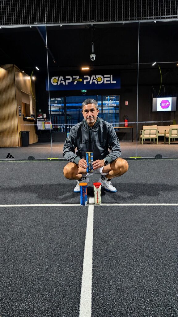Bullpadel | Padel Magazine Man in a gray hoodie crouches on an indoor track, holding two protein shaker bottles with three more bottles on the floor in front.