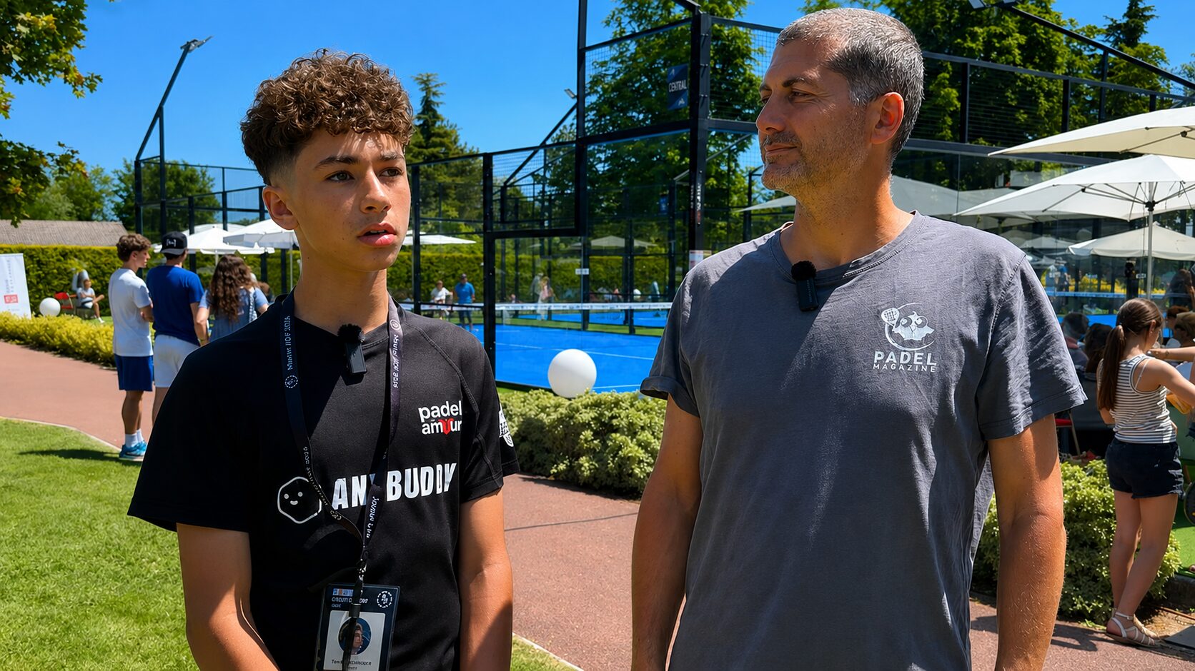 Two men stand near a blue paddle tennis court on a sunny day; the younger wears a black shirt with a lanyard and mic, the older in a gray 'Padel Magazine' shirt listening.