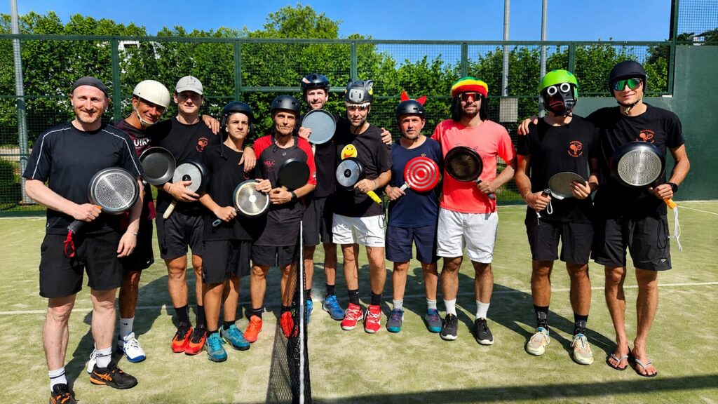 Group of players on a sunny outdoor court posing with frying pans as paddles; some wear helmets and masks, standing in a line behind a net.
