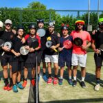 Group of players on a sunny outdoor court posing with frying pans as paddles; some wear helmets and masks, standing in a line behind a net.