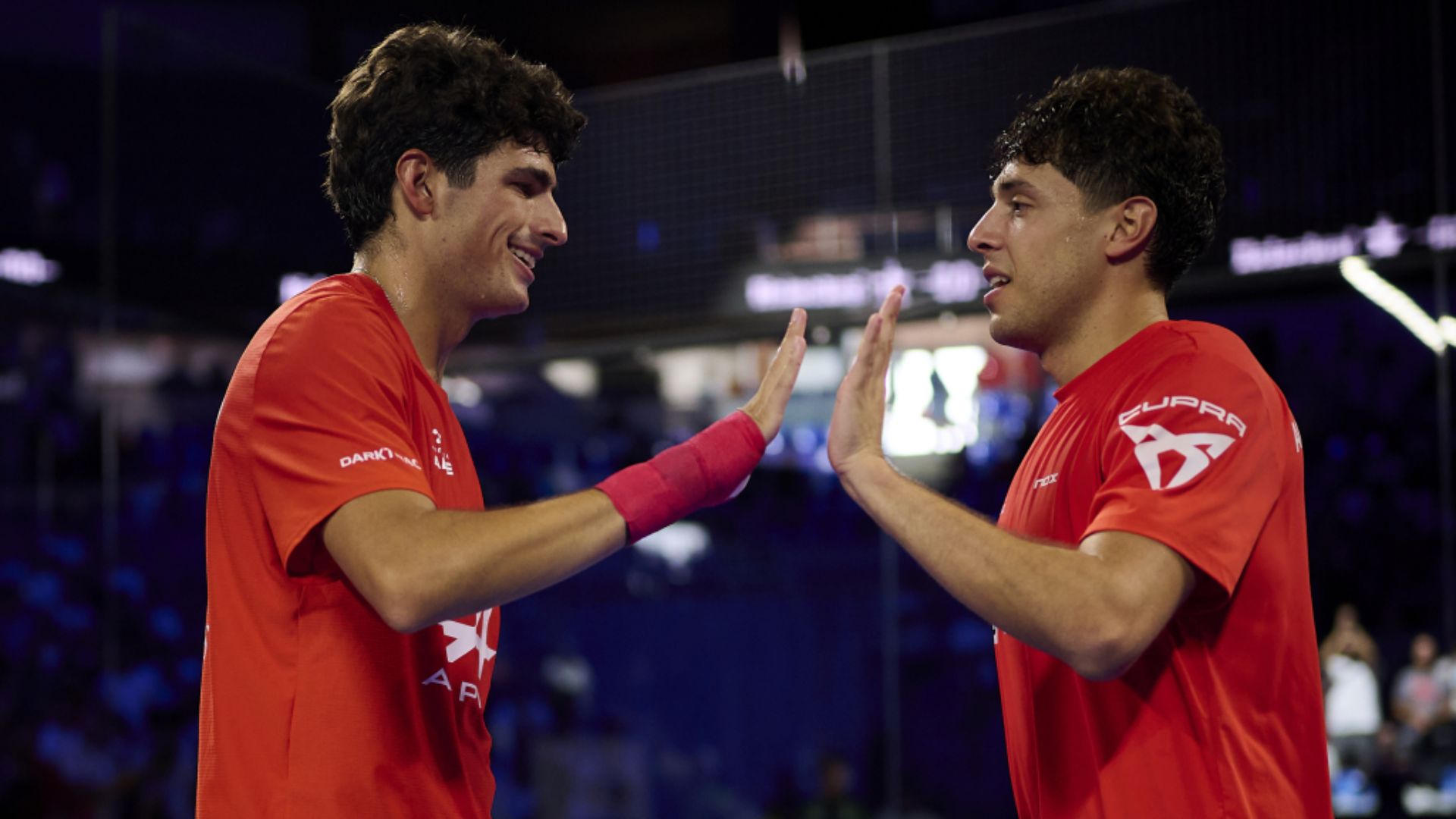 Two male athletes in red jerseys high-fiving in a dimly lit sports arena.
