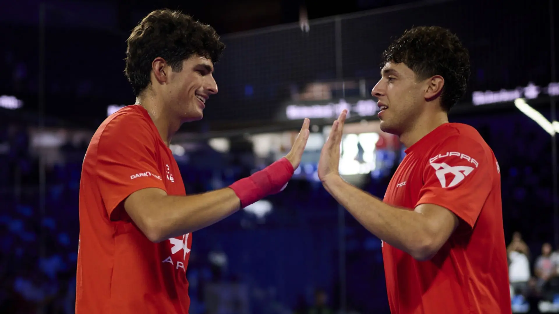 Two male athletes in red jerseys high-fiving in a dimly lit sports arena.