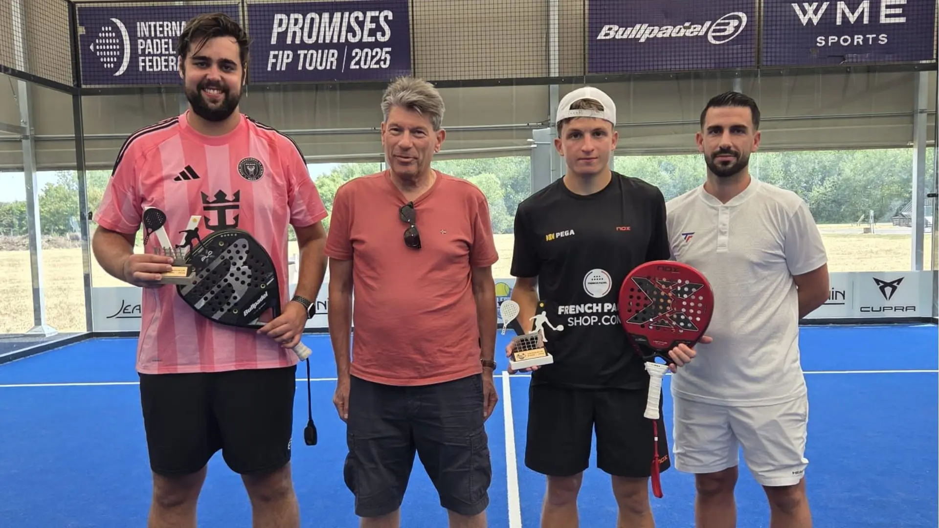 Four men stand on a blue padel court posing with trophies and paddles after a tournament, banners in the background.