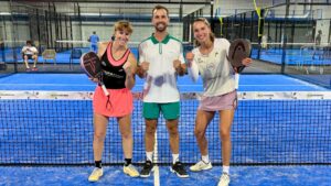Tre padelspelare poserar med paddlar på en blå bana, leende och med nävarna i höjd i firande.