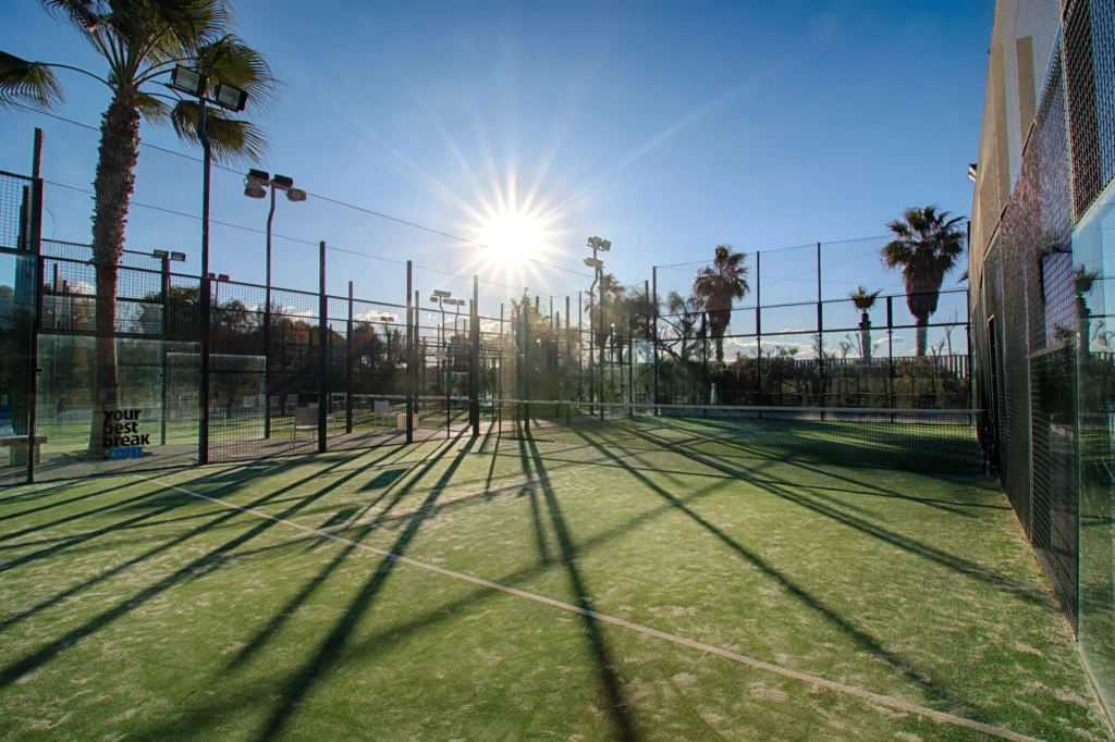 Campo da tennis all'aperto, recintato e illuminato dal sole, con palme e lunghe ombre che si stagliano sulla superficie verde sotto un cielo azzurro e limpido.