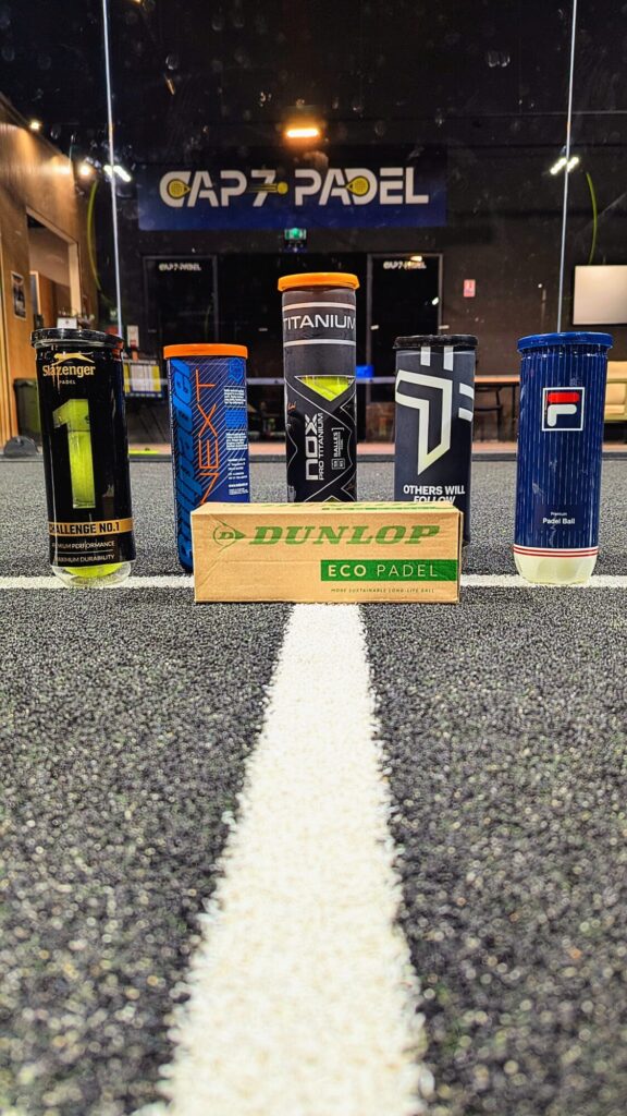 Durables | Padel Magazine Five colorful sport drink cans lined up on a padel court, with a Dunlop Eco Padel box in front and Cap7 Padel signage in the background.