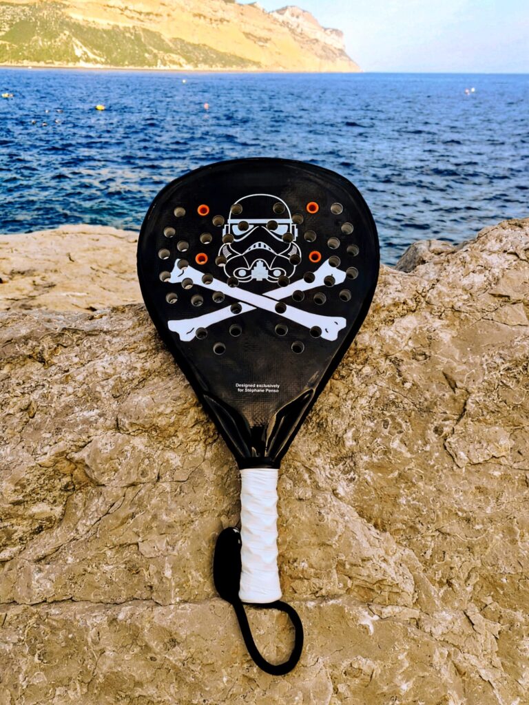 Padel racket resting on a rocky ledge with the blue sea and cliff in the background.