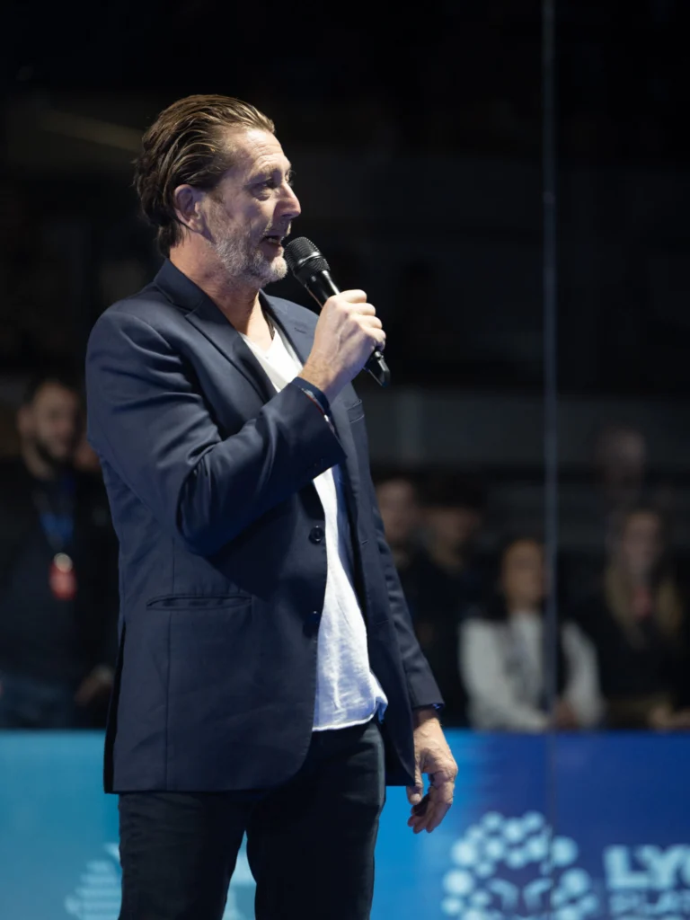 Man in a navy blazer speaks into a handheld microphone on a stage, with an out-of-focus audience in the background.