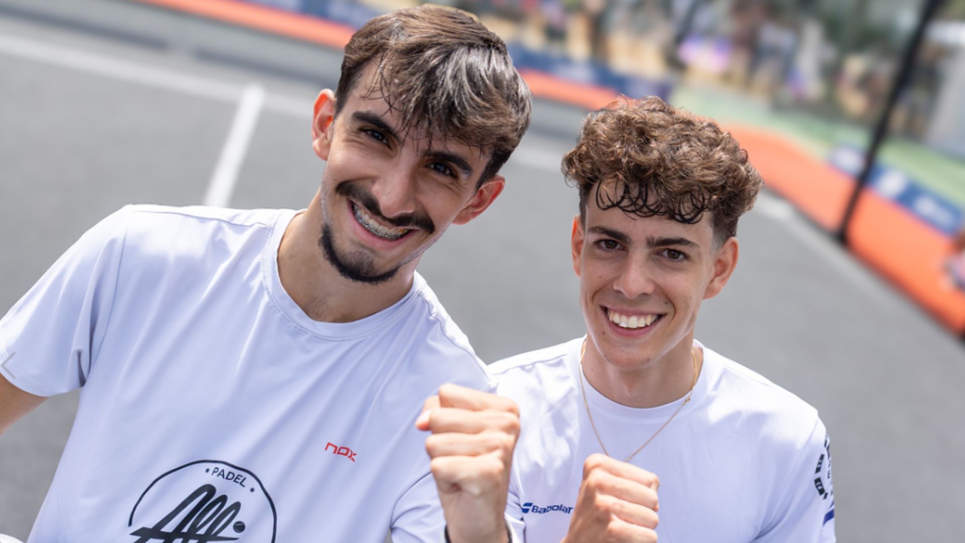 Two young men in white shirts smile at the camera, posing with fists raised in a friendly selfie on an outdoor sports court.