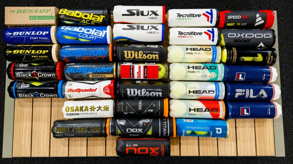 Una fila di manicotti per impugnature da padel impilati su un espositore di legno, che mostrano marchi come Dunlop, Siux, Tecnicafibra, Head, BullpadelWilson e Osaka.