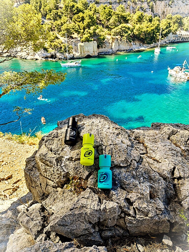 Gel Cassis | Padel Magazine Rocky shoreline overlooking a turquoise sea with anchored boats and green trees in the background; colorful water bottles sit on the rock foreground.