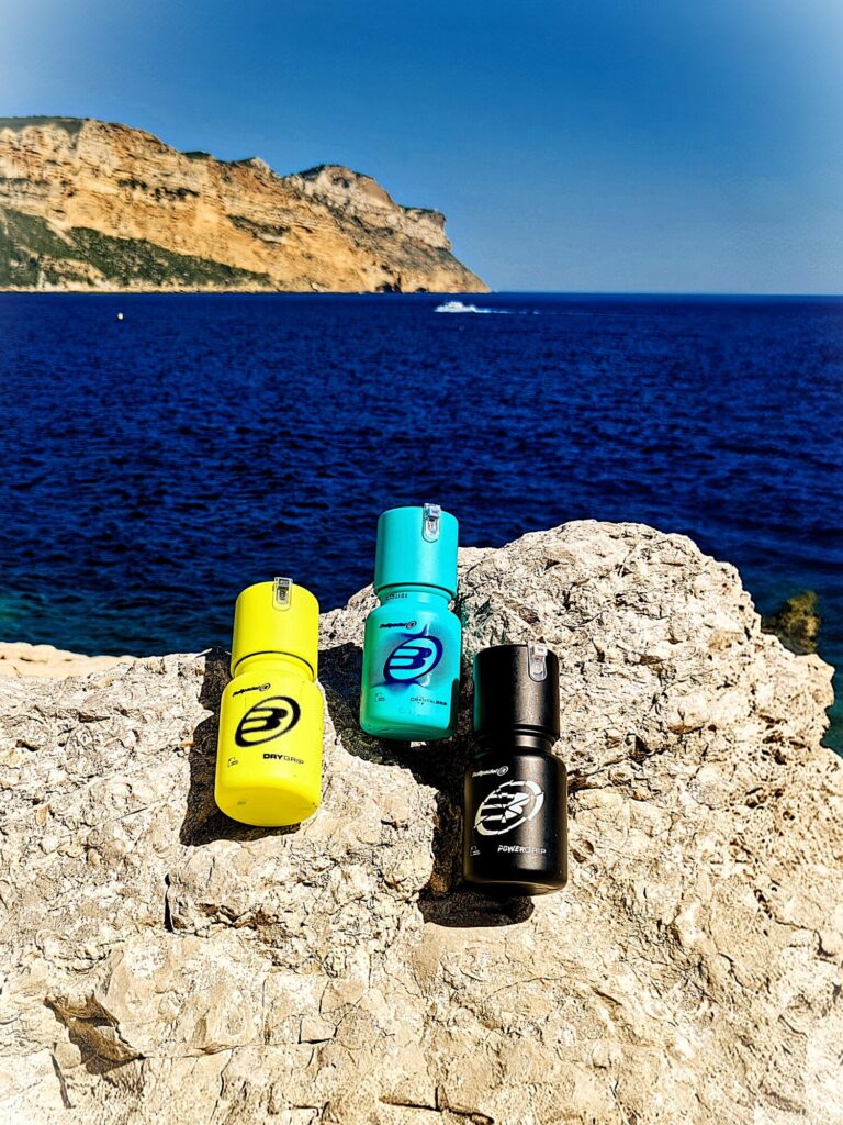 Gel Cassis Bis | Padel Magazine Five colorful sports water bottles arranged on a rocky coastline with the blue sea and cliff in the background.