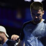 Guichard et Geens butent sur García et Jiménez en 16e du Málaga P1 | Padel Magazine Male esports player with a beard in a blue patterned jersey reading 'Raptor' stands on a dimly lit stage, looking downward still.