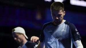 Male esports player with a beard in a blue patterned jersey reading 'Raptor' stands on a dimly lit stage, looking downward still.