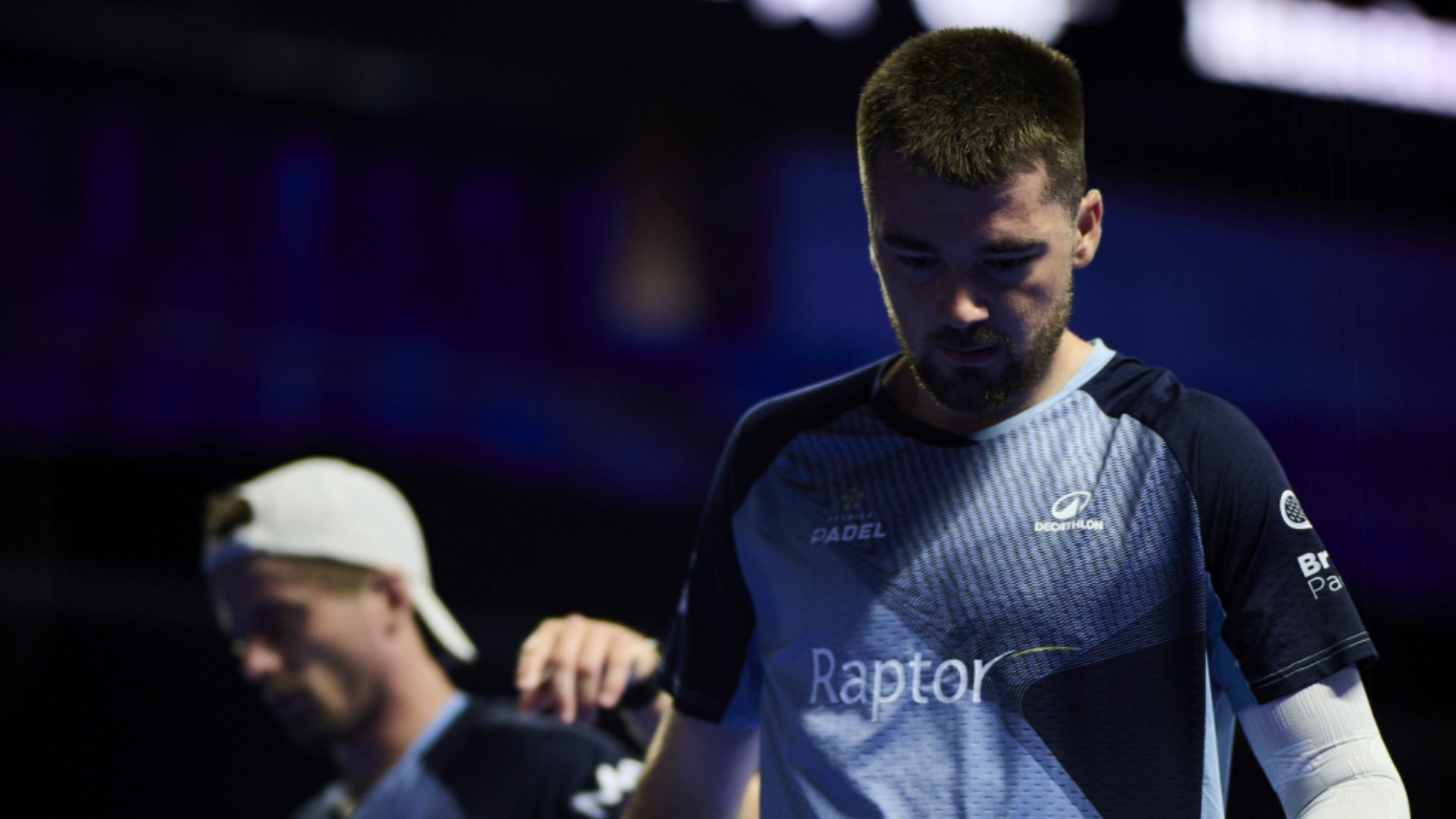 Male esports player with a beard in a blue patterned jersey reading 'Raptor' stands on a dimly lit stage, looking downward still.