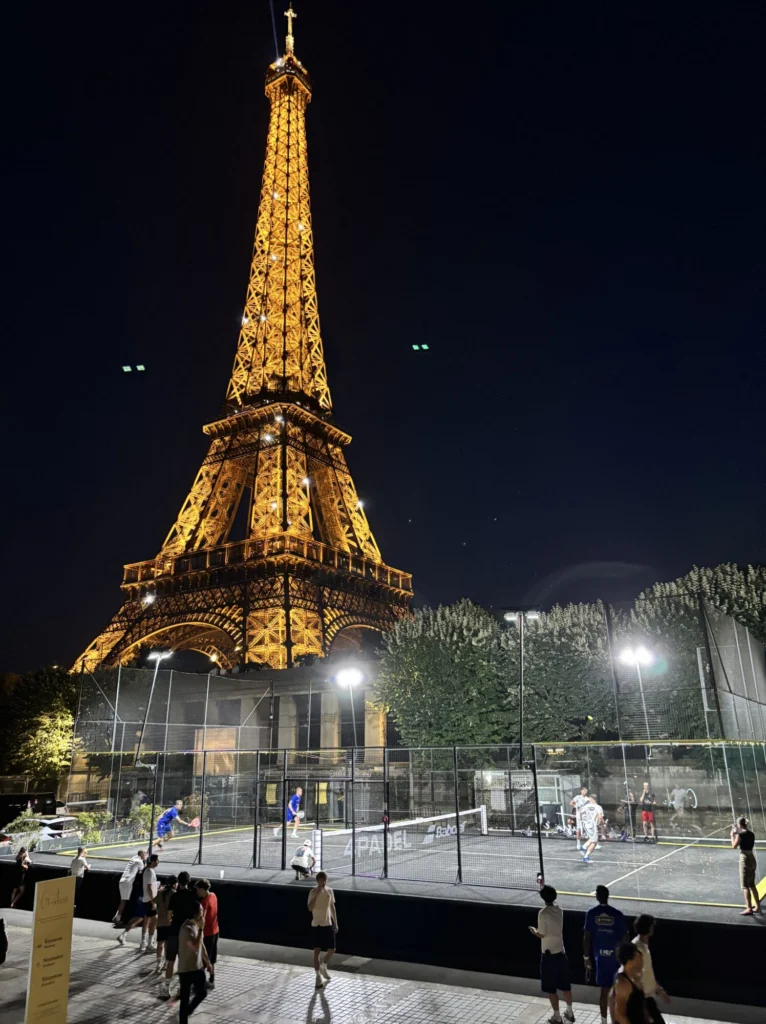Veduta notturna della Torre Eiffel illuminata d'oro, con un campo da tennis all'aperto recintato e i giocatori in primo piano.