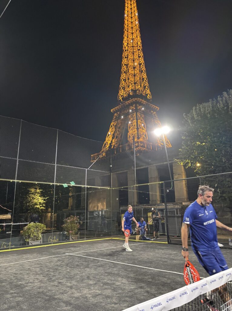 Nighttime padel court with players in blue shirts; an illuminated lattice tower rises in the background.