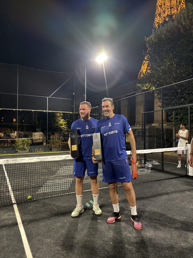 Two men in blue sports jerseys posing with trophies on a nighttime padel court, smiling after a win.