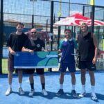 Joule et Boutier créent la surprise et remportent le P1000 Baillargues | Padel Magazine Four men stand side by side on a bright blue padel court, proudly holding a large blue prize check and a trophy after a win, with a fenced court and sponsor tent in the background.