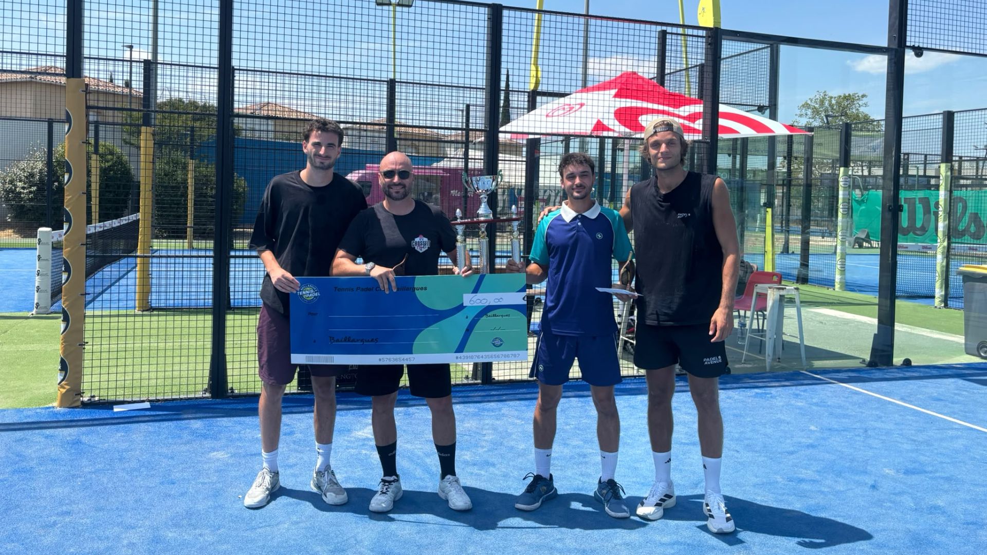 Four men stand side by side on a bright blue padel court, proudly holding a large blue prize check and a trophy after a win, with a fenced court and sponsor tent in the background.