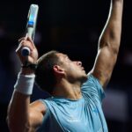 Athlete in a blue shirt raises arms in celebration during a padel match, holding a paddle racket in one hand.