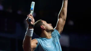 Athlete in a blue shirt raises arms in celebration during a padel match, holding a paddle racket in one hand.