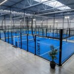 Indoor sports hall with blue paddle (padel) courts enclosed by glass walls and black frames, overhead lighting, and spectator benches along the side.
