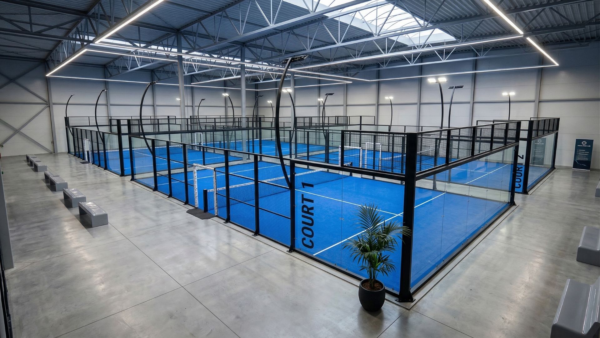 Indoor sports hall with blue paddle (padel) courts enclosed by glass walls and black frames, overhead lighting, and spectator benches along the side.