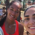 Two smiling women taking a selfie at an outdoor recreation area by a fence, one holding a paddle in the foreground.