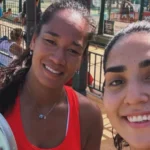 Two smiling women taking a selfie at an outdoor recreation area by a fence, one holding a paddle in the foreground.