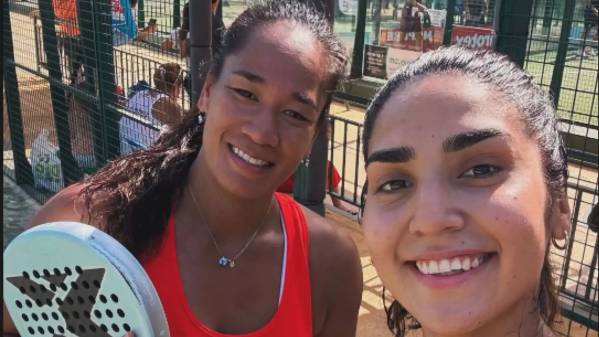 Two smiling women taking a selfie at an outdoor recreation area by a fence, one holding a paddle in the foreground.