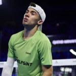 Leo Augsburger Je fais un énorme travail psychologique pour supporter tout cela à mon âge | Padel Magazine Squash player in a lime green shirt and white cap, standing near the net in a dim arena, looking upward.