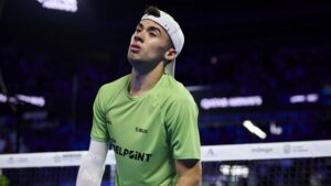 Squash player in a lime green shirt and white cap, standing near the net in a dim arena, looking upward.
