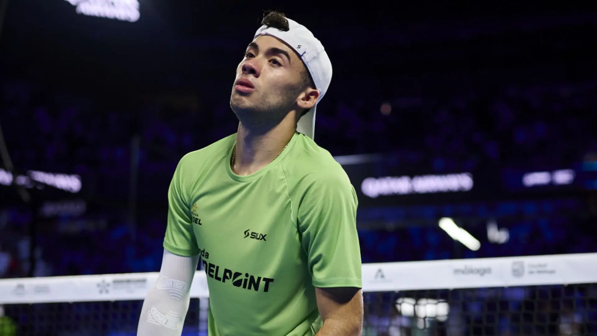 Squash player in a lime green shirt and white cap, standing near the net in a dim arena, looking upward.