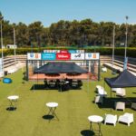 Outdoor event setup on a tennis court with white chairs, round tables, and black and red tents beneath a clear blue sky, sponsor banners along the fence.