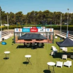Outdoor event setup on a tennis court with white chairs, round tables, and black and red tents beneath a clear blue sky, sponsor banners along the fence.