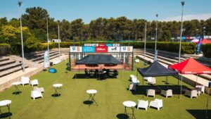 Outdoor event setup on a tennis court with white chairs, round tables, and black and red tents beneath a clear blue sky, sponsor banners along the fence.