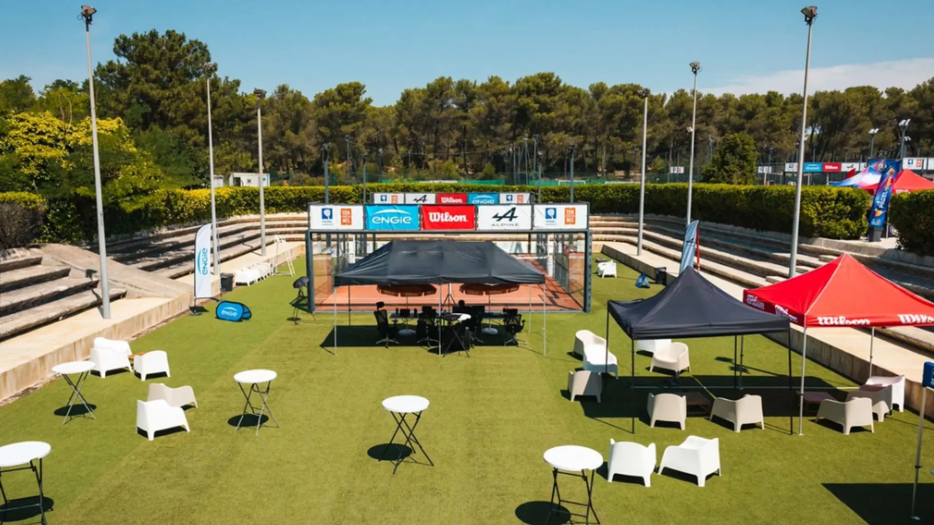 Outdoor event setup on a tennis court with white chairs, round tables, and black and red tents beneath a clear blue sky, sponsor banners along the fence.