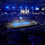 Où voir les finales du Premier Padel Málaga P1 | Padel Magazine Indoor arena packed with spectators watching a tennis match on a bright blue court under blue lights, with a large scoreboard overhead and screens around the arena.