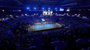 Indoor arena packed with spectators watching a tennis match on a bright blue court under blue lights, with a large scoreboard overhead and screens around the arena.