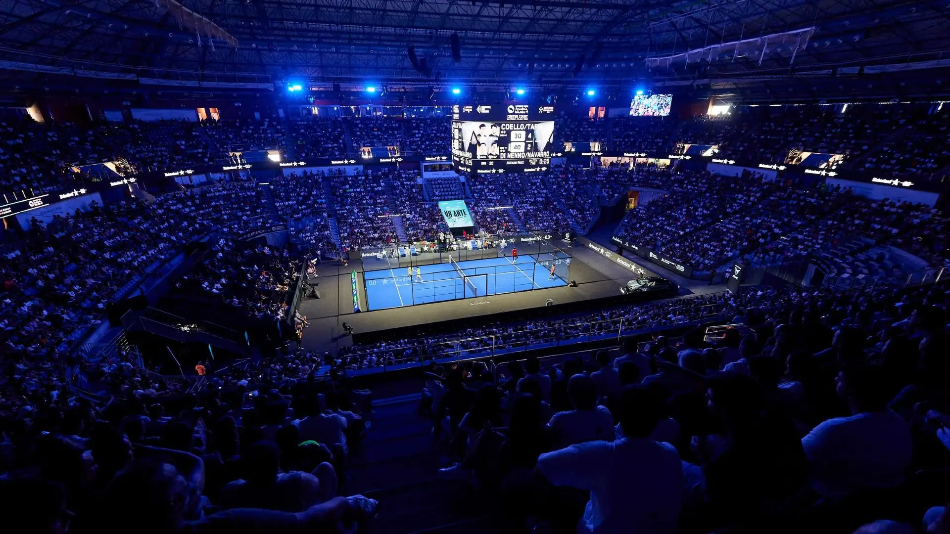Indoor arena packed with spectators watching a tennis match on a bright blue court under blue lights, with a large scoreboard overhead and screens around the arena.